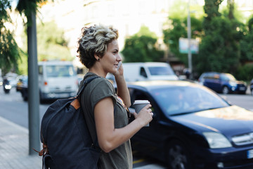 Blonde tomboy girl is talking by a mobile phone while standing with a blank paper cup of coffee on a blurred urban background. Student female is chatting with a friend by a smartphone.