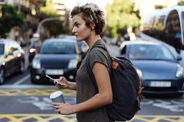 Happy young woman with blonde short hair is messaging to the friend by a smartphone while crossing...