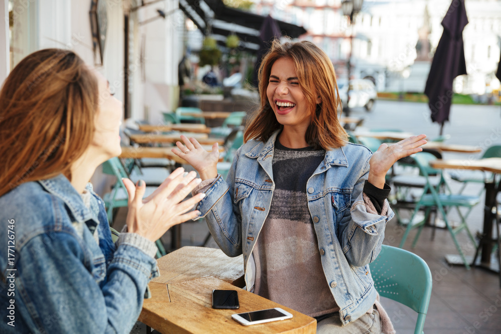 Poster two emotional young girls talking while sitting in city cafe