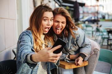 Close-up portrait of funny young woman showing tongue while taking selfie on mobile phone, city outdoor