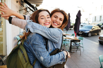 Two young happy brunette girls hugging each other, looking at camera, city outdoor