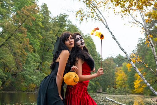 Two young brunettes women with makeup like a Halloween skull and Halloween witch makeup stands with a torch in a red and black dresses