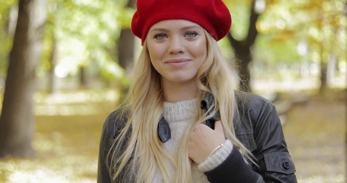 Attractive young woman in leather jacket and red beret standing on alley, smiling and looking at camera during her walk in autumn park.