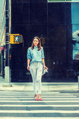 Fototapeta na wymiar Young Beautiful Woman wearing light blue striped shirt, white destroyed jeans, red sandal heels, shoulder carrying small bag, walking across street by traffic light in New York. .