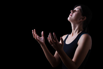 Low key of a faithful woman praying and feeling the presence or being touched by god. Arms outstretched in worship, head up and eyes closed in religious fervor. Black background. - obrazy, fototapety, plakaty