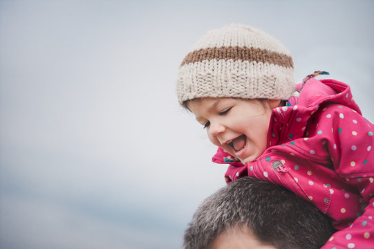 Cute little girl wearing jacket and hat sitting on her father's shoulder and laughing.
