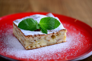 A piece of apple pie, sprinkled with sugar, adorned with a sprig of fresh mint. Served on a red plate, close-up.