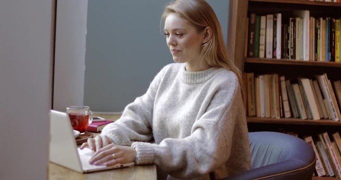 Beautiful young woman in sort jumper sitting in comfortable armchair near bookcase, using laptop and enjoying silence.