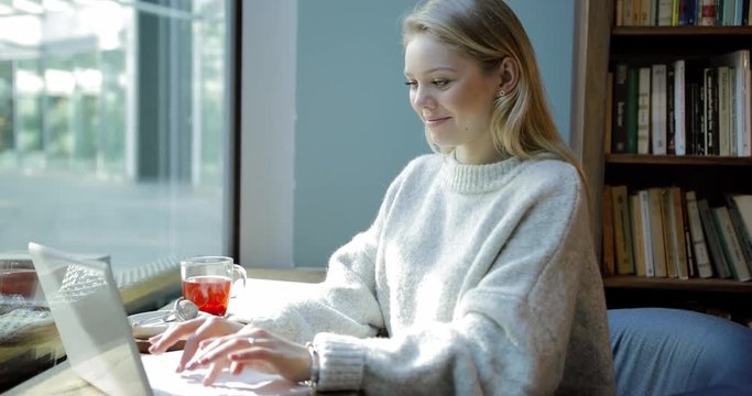 Pretty young woman in soft beige jumper sitting near window with mug of tea and using her laptop.