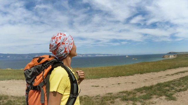 Hiking woman with backpack walking along the road near the blue lake Baikal. Traveler girl on a hiking trail, travel and active lifestyle concept