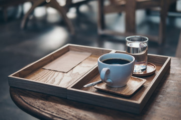 Closeup image of a cup of hot coffee and a glass of water in vintage wooden tray on the table in...