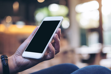 Mockup image of woman's hands holding white mobile phone with blank black screen on thigh in cafe
