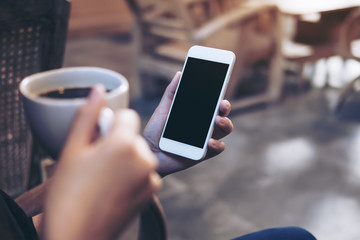Mockup image of woman's hands holding white mobile phone with blank black screen while drinking coffee in modern loft cafe