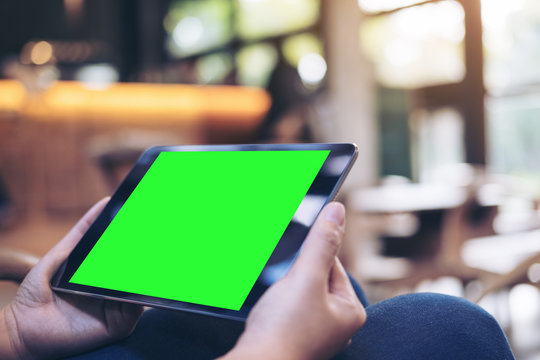 Mockup image of woman's hands holding black tablet pc with blank green screen on thigh with concrete floor background in modern cafe