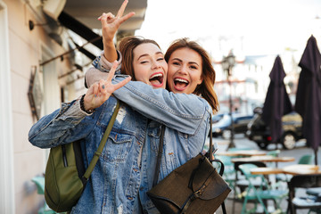 Photo of two charming brunette female friends hugging each other, showing peace sign, looking at camera on city street
