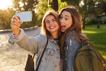 Close-up photo of two funny girls in casual jeans wear making selfie on smartphone, outdoor