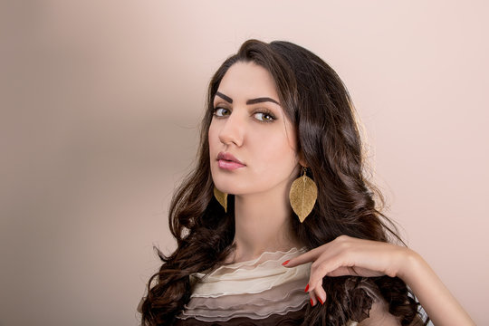 Portrait Of Beautiful Young Mediterranean And Middle Eastern Brunette Hair Girl With Leaf Shape Gold Earrings, Glossy Lips Make Up And Red Nails. Studio Shot On Beige Background