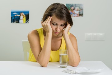 An adult female suffers from a severe headache. She holds her head in her hands. On the table a glass of water and pills. Age, medicine, health care and people concept