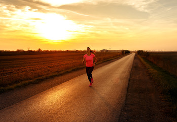 Obraz premium Athletic woman running on rural road during sunset.