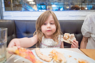 Three years old blonde girl with white dress, taking pizza portion with hand, sitting in black couch next to mother in restaurant  
