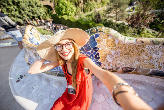 Young woman tourist in red dress making selfie photo sitting on the bench decorated with mosaic in the famous Guell park in Barcelona