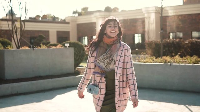charming young woman walks through the autumn city in a coat and smiles. cute girl on a background of modern architecture. slow motion