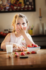 Pretty little girl eating strawberries and jam in a kitchen