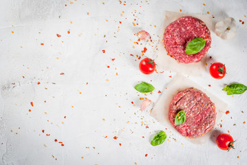 Fresh raw home-made minced beef steak burger with spices, tomatoes and basil, on a white stone concrete table, copy space, top view