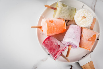 Healthy summer desserts. Ice cream popsicles. Frozen tropical juices, smoothies blueberries. currants, orange, mango, kiwi, banana, coconut, raspberry. On white marble table, plate copy space top view