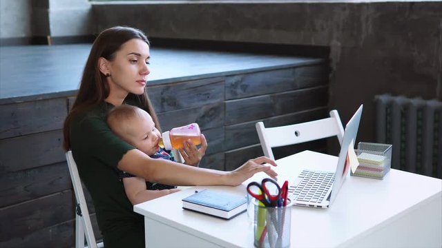 Woman with dark hair is watering her little child from a bottle. She is sitting in a modern room at a table, laptop, diary and stickers are lying on a table.