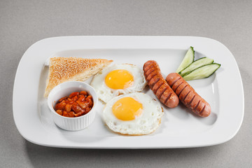 Fried eggs with sausages, beans, cucumber and bread on white plate