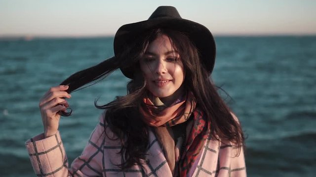 portrait of cute girl closeup on sea background. young woman in autumn coat and hat posing and smiling at the camera.. Slow motion
