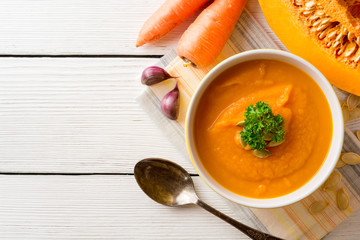 Pumpkin and carrot cream soup with pumpkin seeds and parsley in bowl on white wooden background.