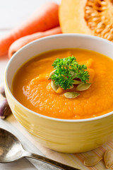 Pumpkin and carrot cream soup with pumpkin seeds and parsley in bowl on white wooden background.