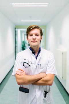 Friendly young doctor standing in the hall of a hospital with his arms crossed