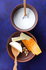 Camembert cheese, orange cheese, peanuts, wooden spoon and spatula with clay ware on a dark blue background