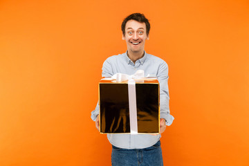 Mimicry and crazy concept. Young adult happiness man, looking at camera with funny rabbit face, and big eyes, holding big gold box with white bow knot.