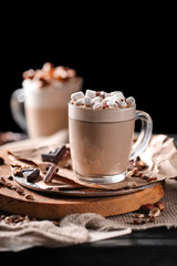 Composition with two glass of cappuccino topped with marshmallow and served with coffee beans and chocolate on wooden chopping board over black background
