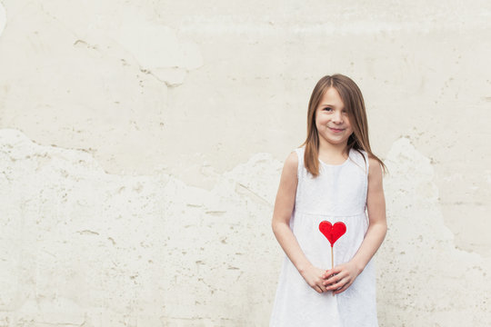 Little Girl In White Dress Holding A Red Candy Heart Lollipop