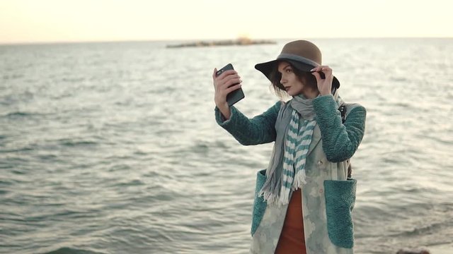 girl doing selfie on the beach at sunset. young attractive woman in autumn coat takes a picture of herself on a mobile phone camera. slow motion