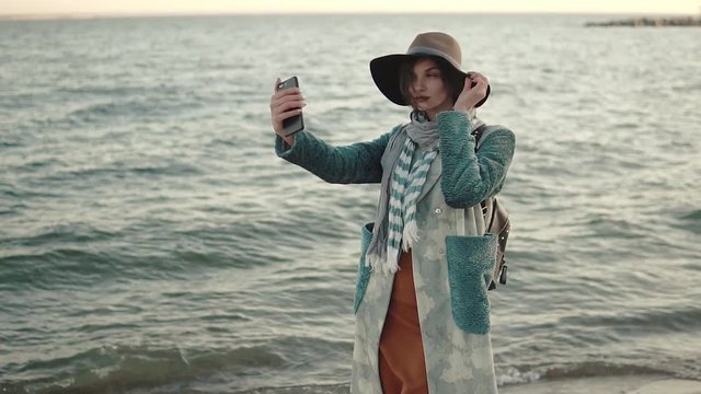 girl doing selfie on the beach at sunset. young attractive woman in autumn coat takes a picture of herself on a mobile phone camera. slow motion