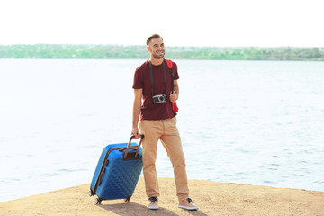 Handsome young tourist with suitcase standing on river bank