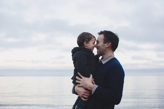 Father and son rubbing noses by sea on overcast day