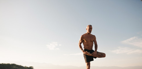 Muscular, fit senior man doing yoga (tree pose) outside in morning sunlight