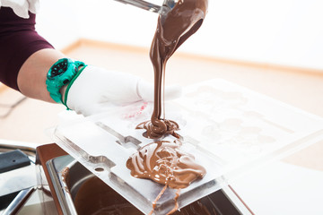 A close-up of a woman with a conditor in a white apron pours a hot spoonful of hot milk chocolate over a large spoon from a large pot. Preparation of chocolate sweets - Powered by Adobe