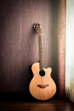 Acoustic guitar vintage style lean on the wall in low light corner. Soft light from the window provides some details on the old guitar