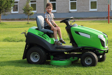 Little boy sits on lawn on green grass near building at summer day