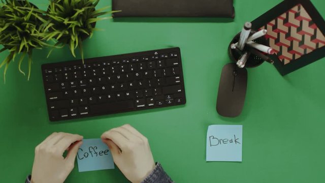 Top down view of man sticking notes with words "coffee break" on them and putting cup of coffee on green table between them