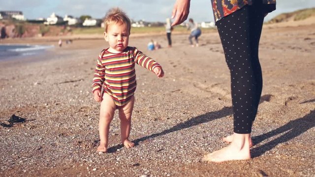 A little baby boy is playing and learning to walk with his mother on the beach at sunset