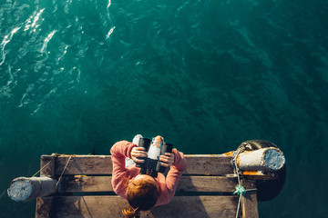 Young Girl Sits On The Pier And Looks At Sea Through Binoculars, Top View. Adventure Vacation...